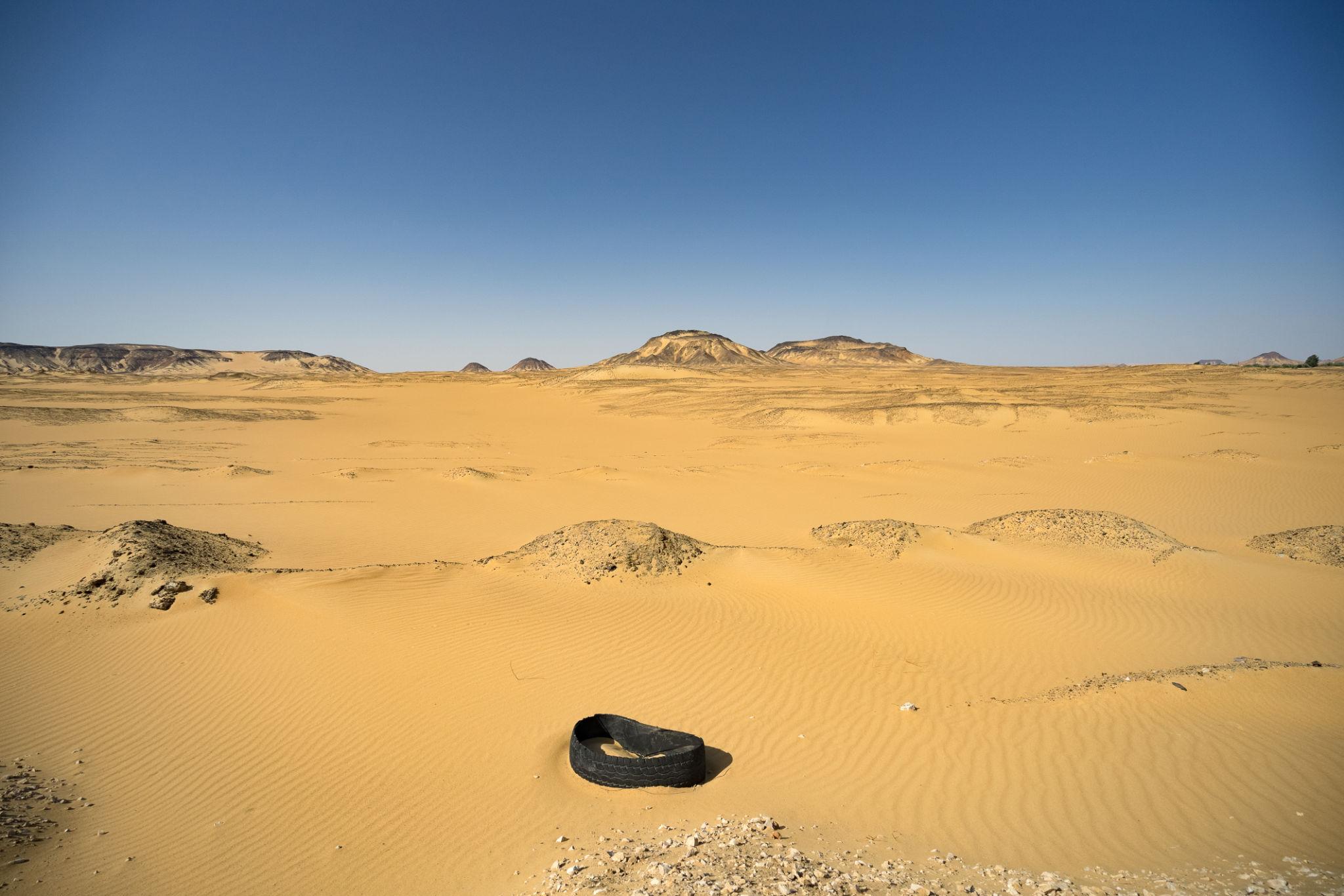 A discarded tire half-buried in rippled sand dunes with rocky hills in the distance
